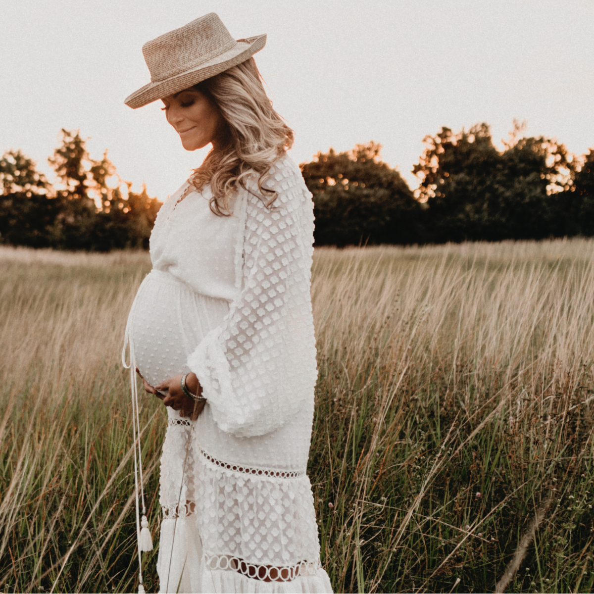 Pregnant Person In White Dress In A Field Wearing a Straw Hat